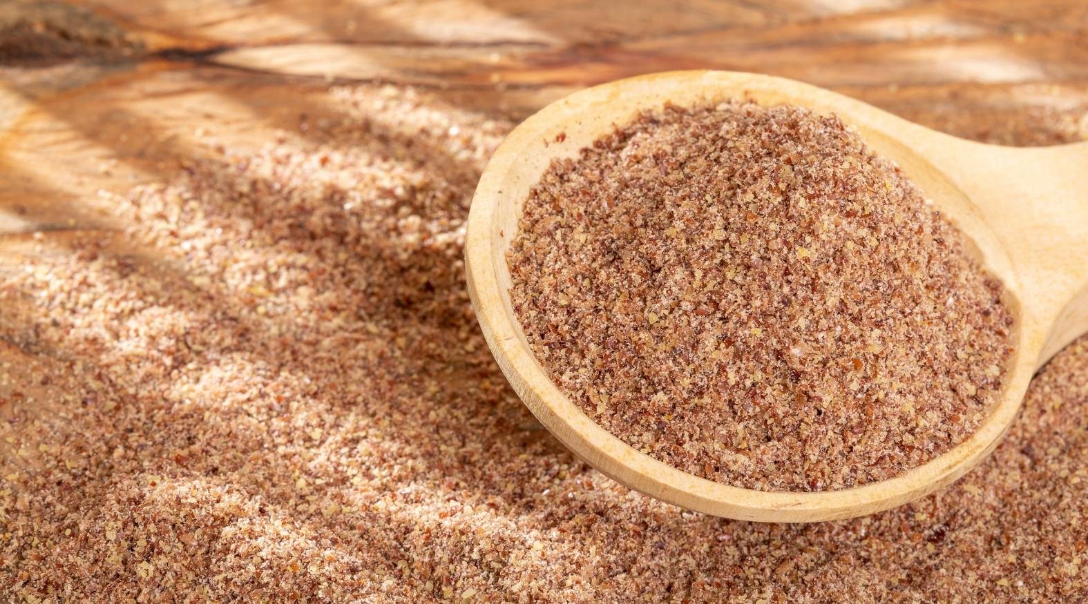 Ground flaxseed (Linum usitatissimum) shown in a wooden spoon, highlighting its fine texture and natural brown colour.