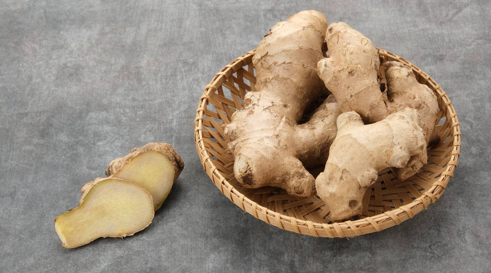Fresh ginger (Zingiber officinale) roots displayed in a woven basket, with sliced pieces showing the pale yellow interior and fibrous texture.