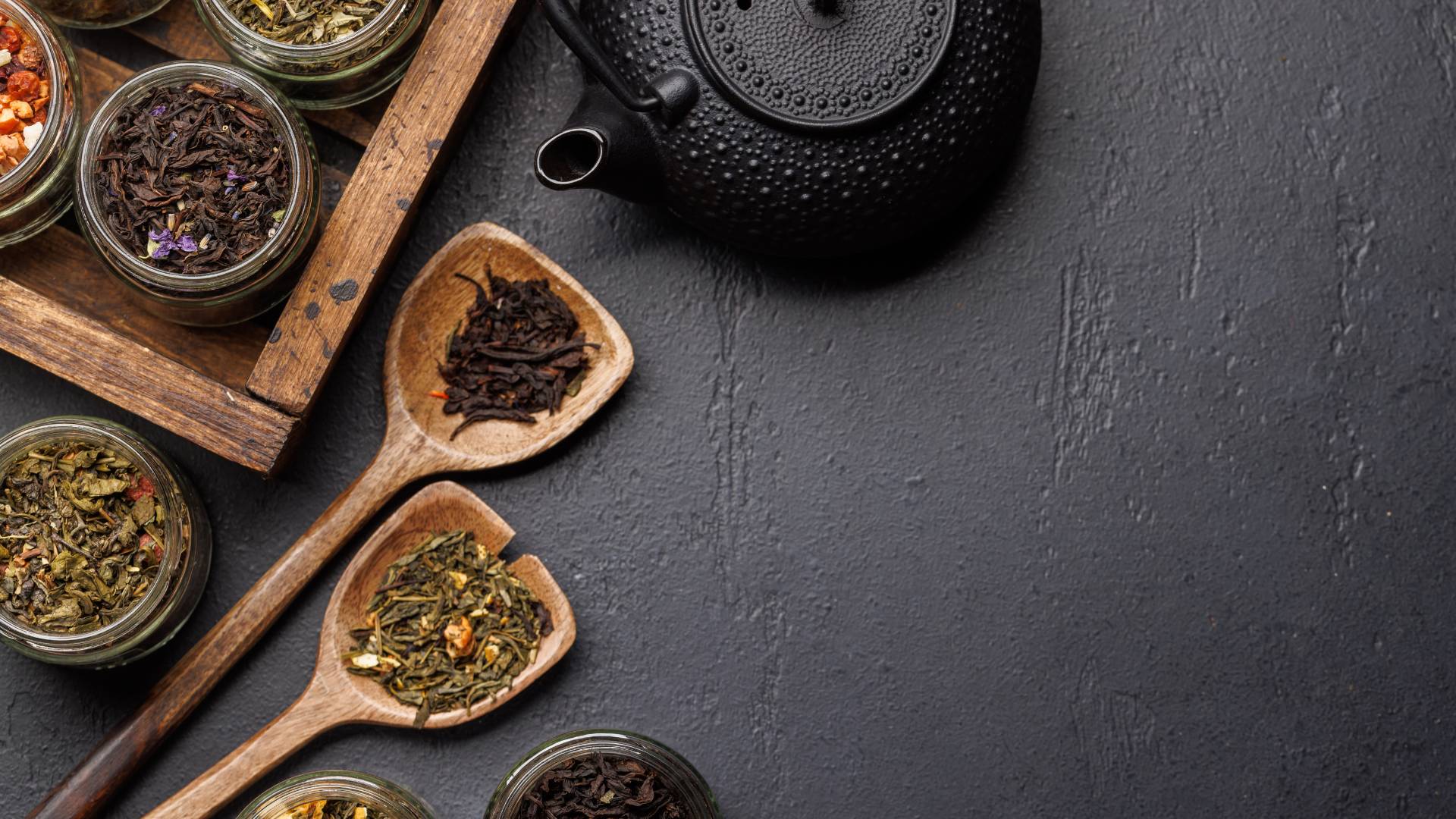 Black teapot with jars and wooden spoons of dried herbs on dark surface, natural herbal tea for energy support