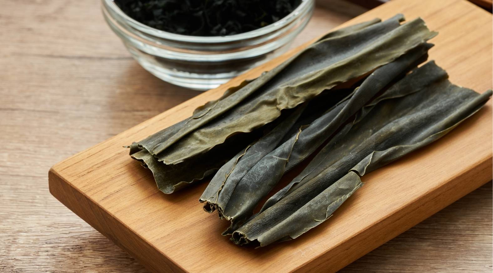 Dried kelp (Laminaria spp.) strips laid on a wooden board, showing their long, flat, slightly wrinkled texture with a muted green-brown colour.