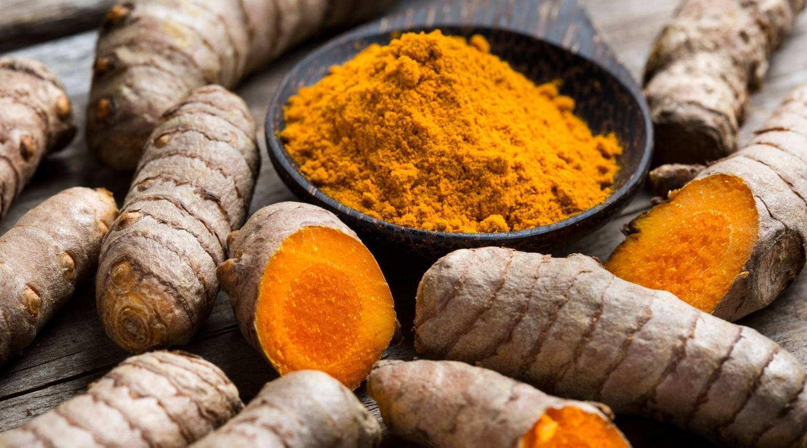 Turmeric (Curcuma longa) featuring fresh rhizomes with textured, earthy brown skin and vivid orange interiors. At the center, a bowl of finely ground turmeric powder displays its rich golden-yellow colour