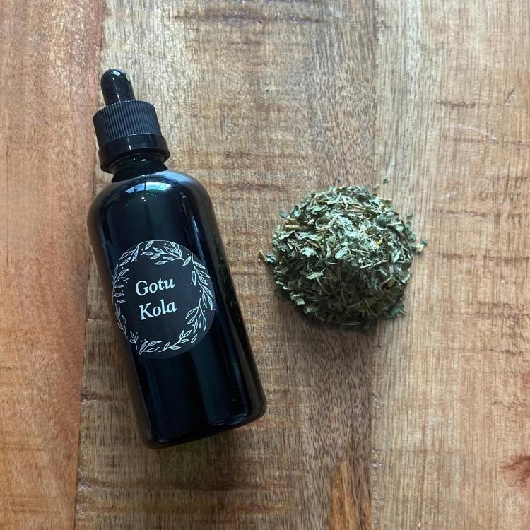 Black bottle labeled 'Gotu Kola' next to a ball of dried herbs on a wooden surface
