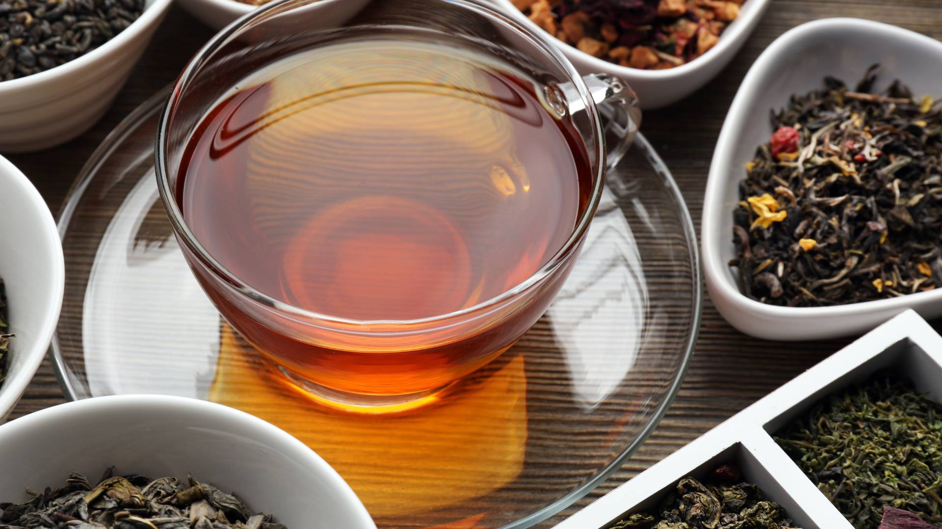 Glass teacup filled with tea surrounded by bowls of different types of tea leaves on a wooden surface.